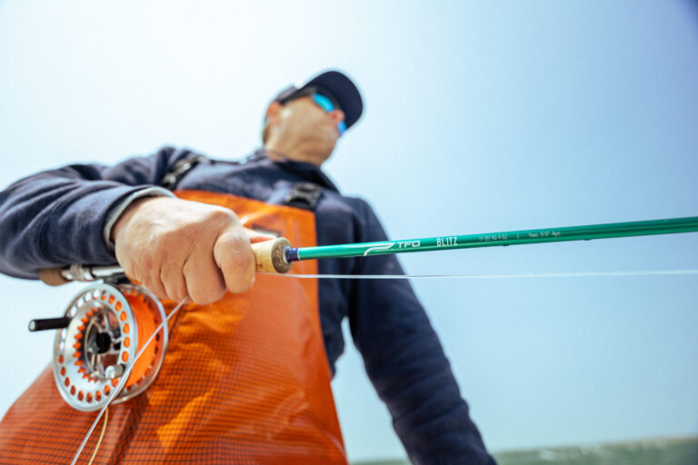 Person holding a fishing rod with an orange vest and blue cap against a clear sky.