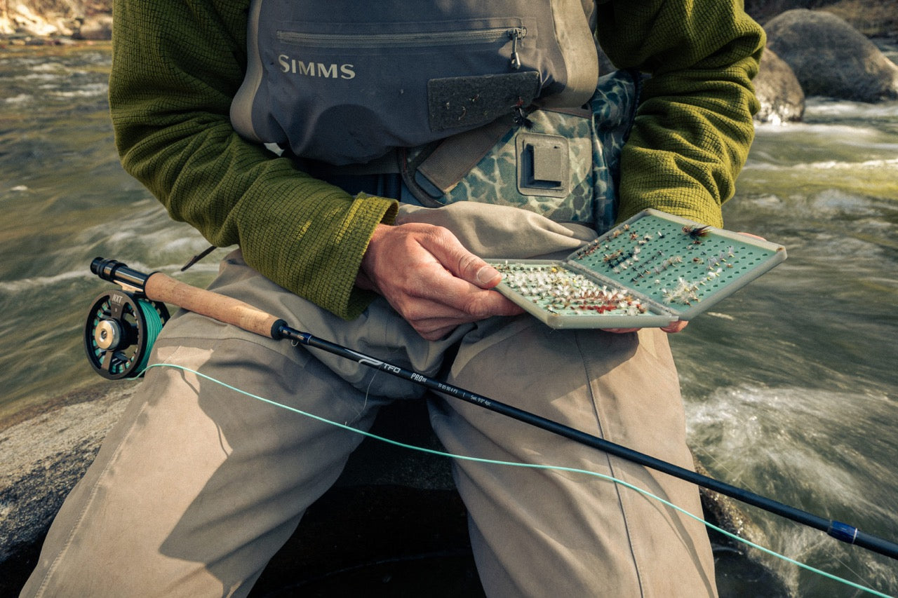 Person holding a fishing rod and a small box with fishing lures by a river.